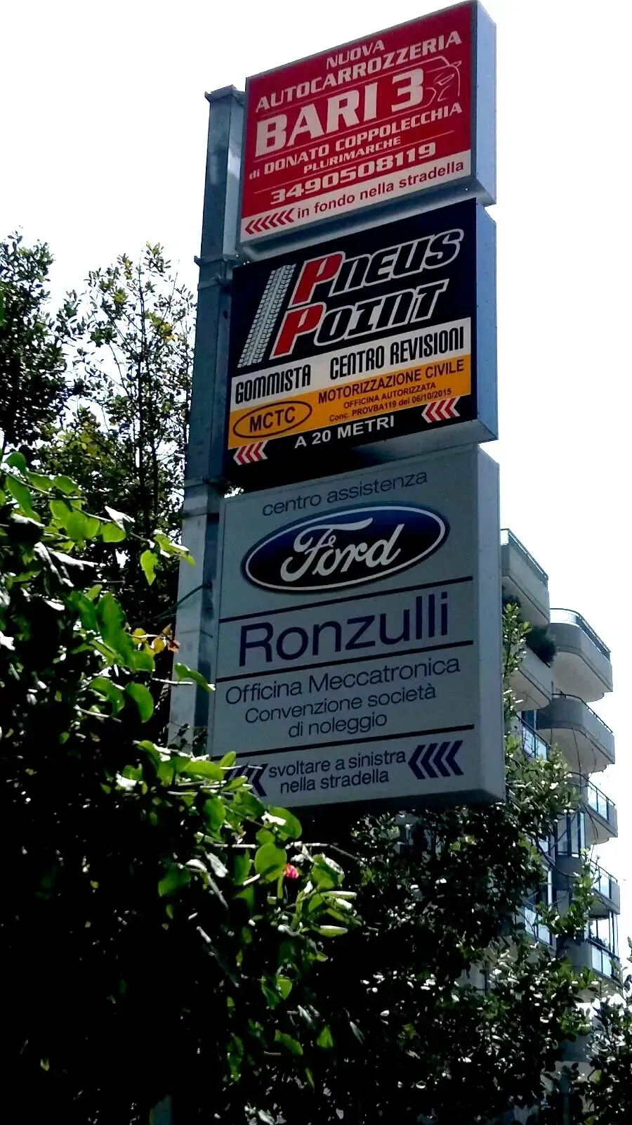Un alto cartellone pubblicitario a bordo strada, con diverse inserzioni, tra cui quella di Ronzulli, si erge al di sopra degli alberi verdi, stagliandosi contro il cielo.