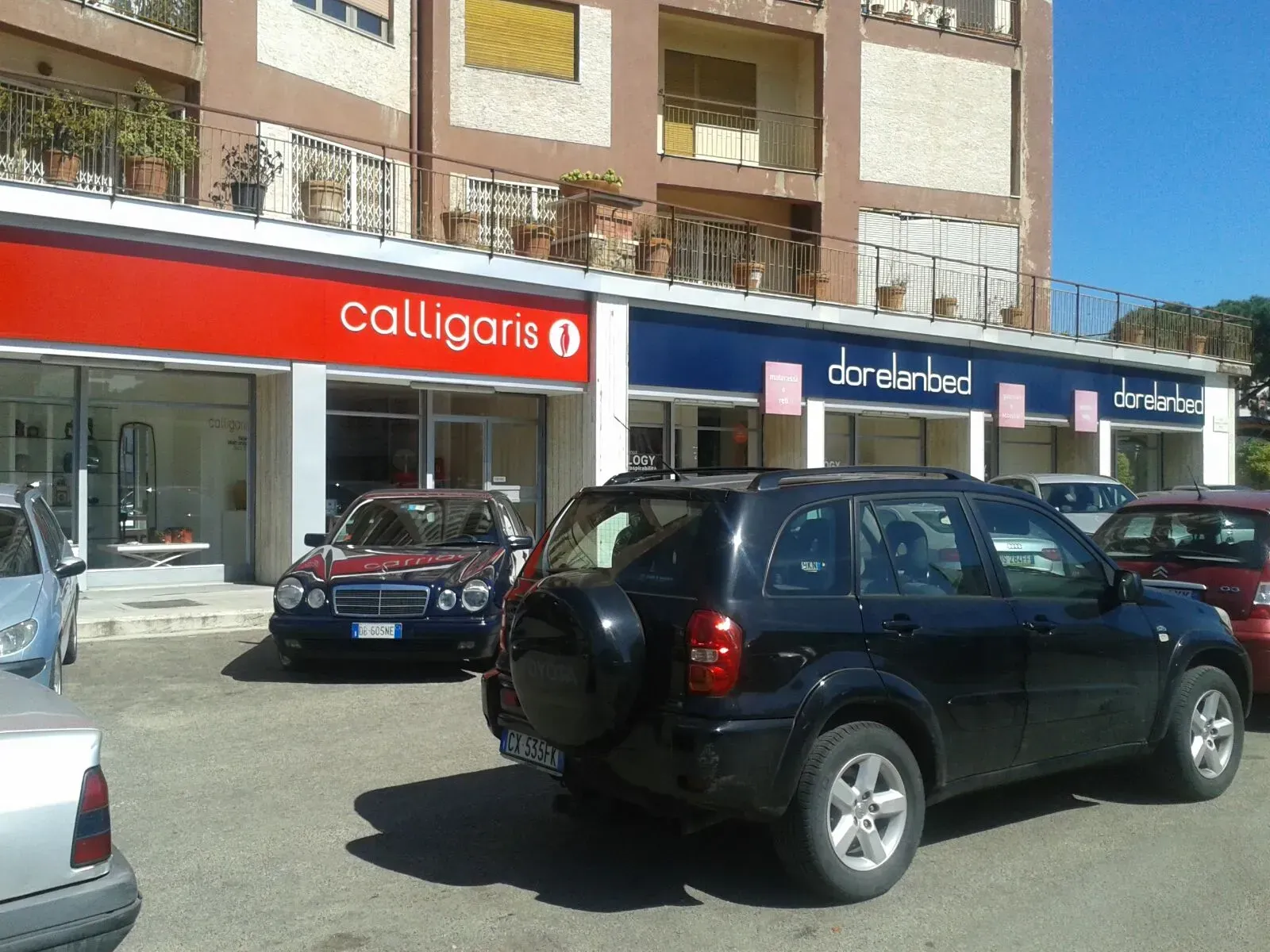 Auto parcheggiate davanti alle vetrine dei negozi con insegne rosse e blu su una strada soleggiata