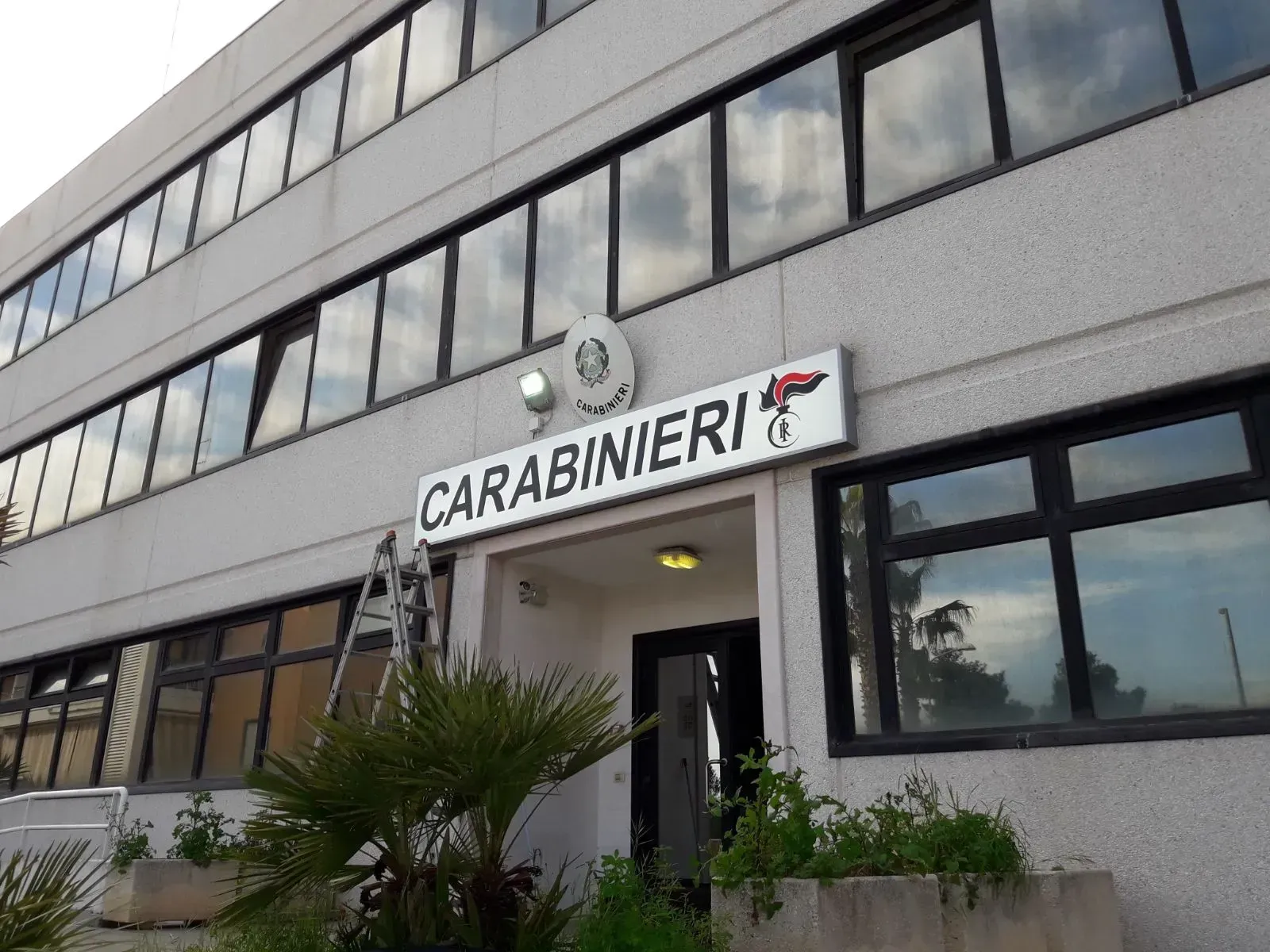 Ingresso della stazione dei Carabinieri su un edificio grigio, con insegna sopra la porta e finestre lungo la facciata.