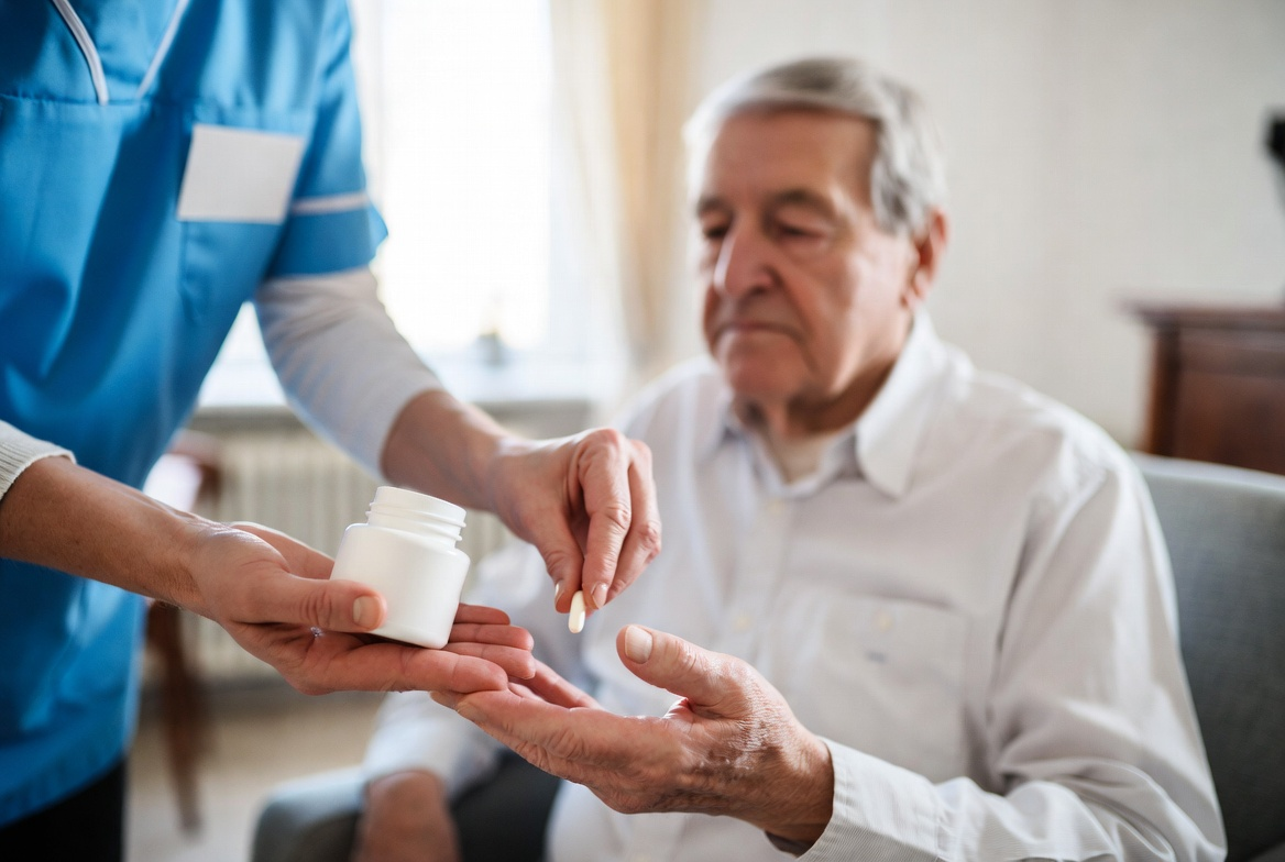 Caregiver giving a pill to a person, indoors. Hands holding pill and white bottle. Elderly man sits, looking at the pill.