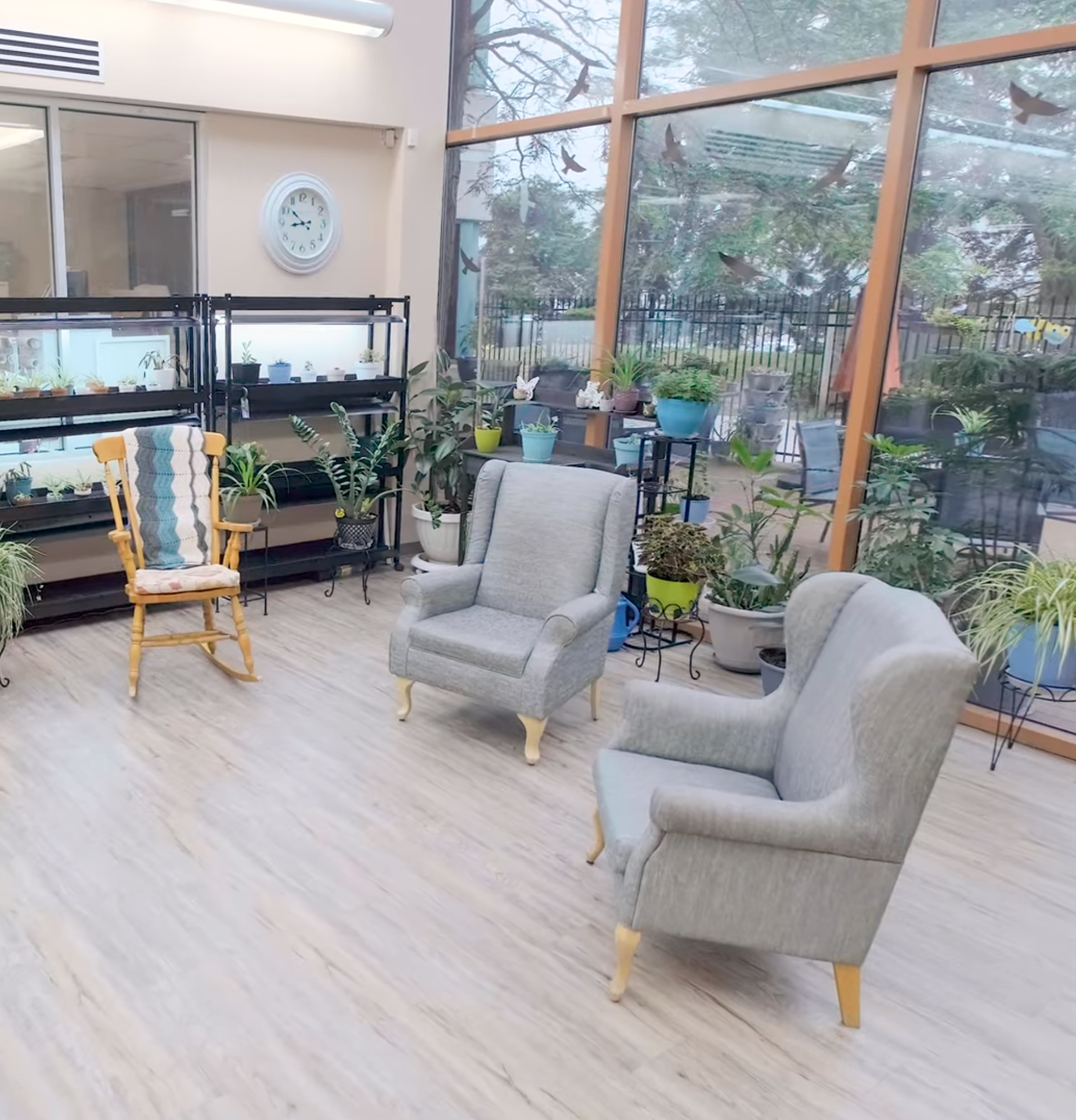 Sunroom with two armchairs, a rocking chair, and plants. Light wood flooring. Large glass windows and beige walls.