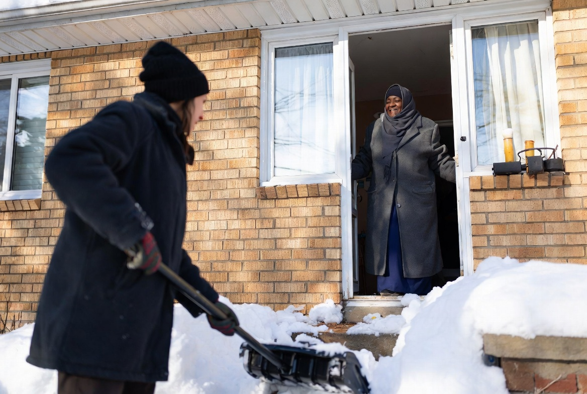 Person shovels snow in front of a brick building; another person stands in the doorway.