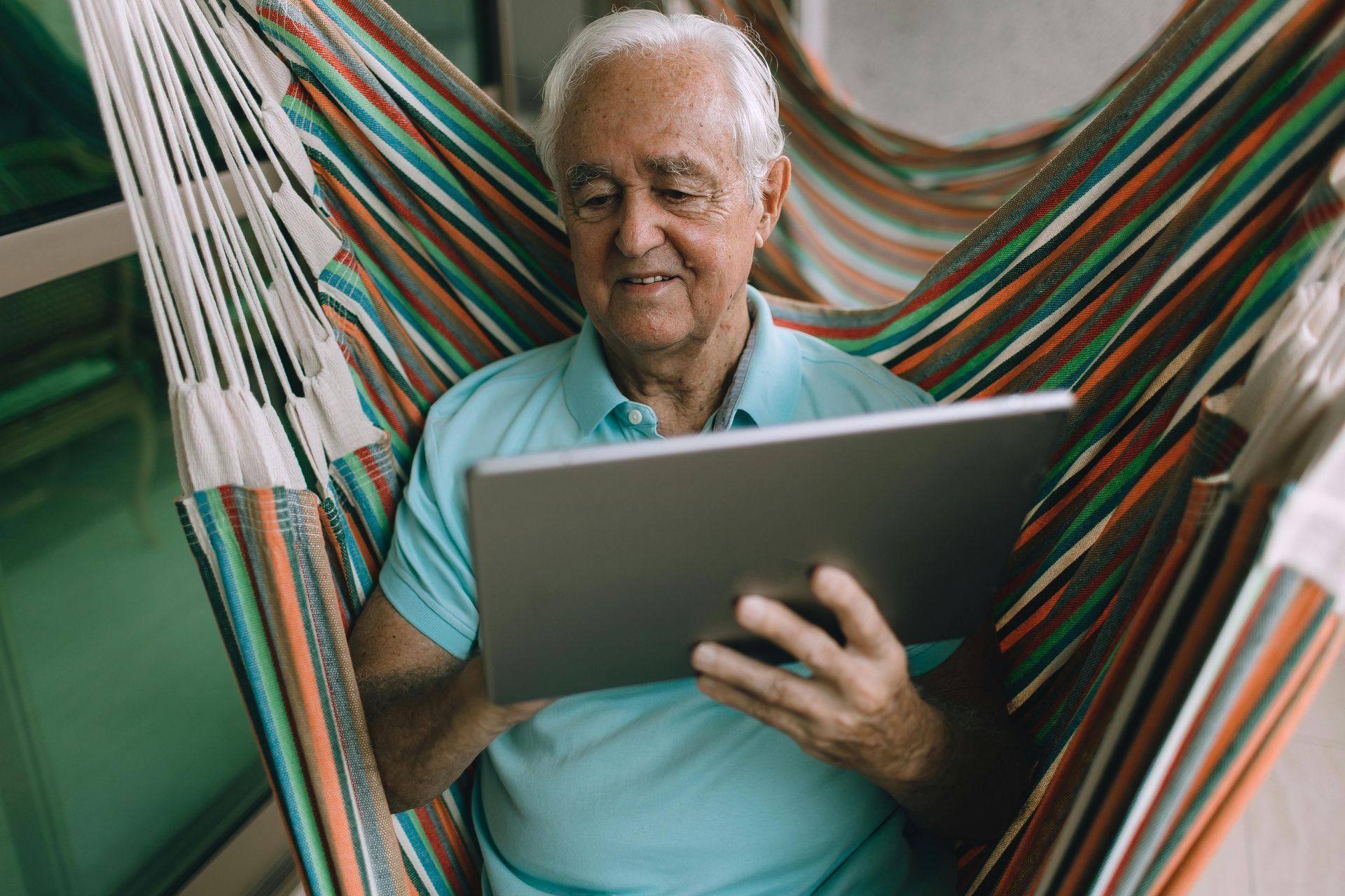 Man in light blue shirt using tablet, reclining in striped hammock.