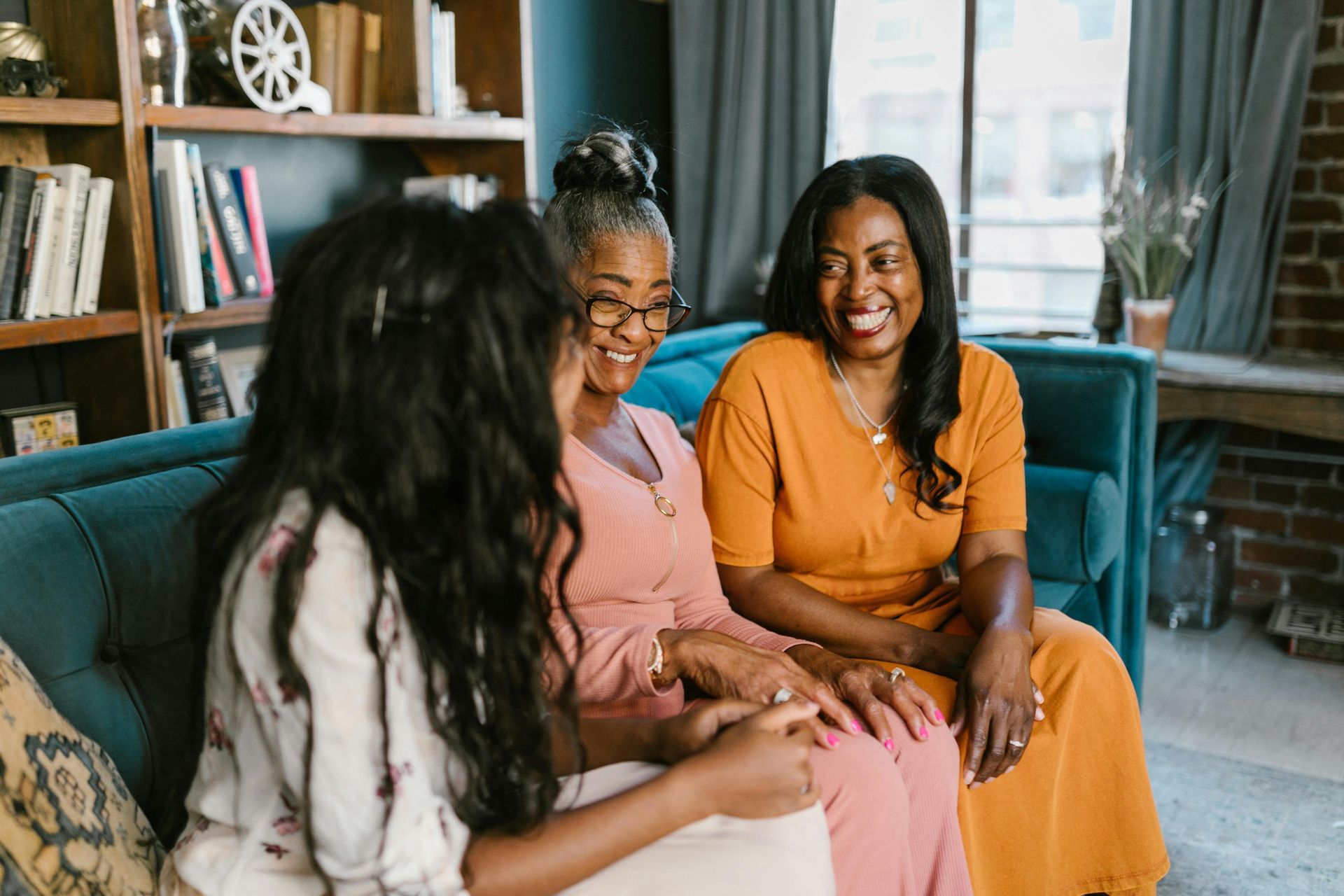Three women smiling and talking on a teal couch in a living room with a bookshelf.