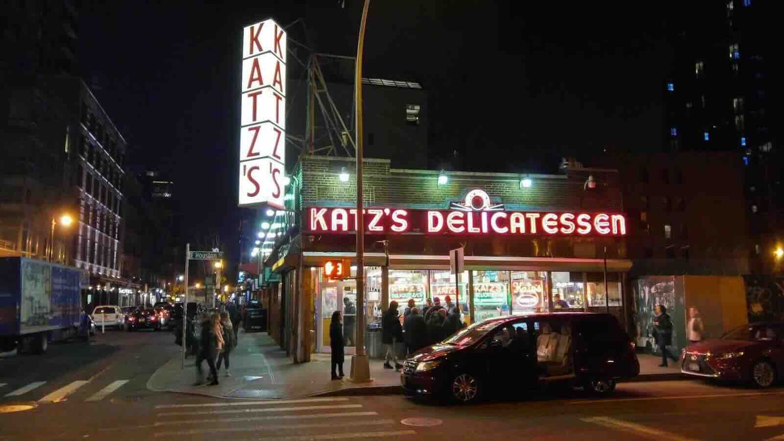 Katz's Delicatessen at night, glowing red sign, people on sidewalk, cars on street in NYC.