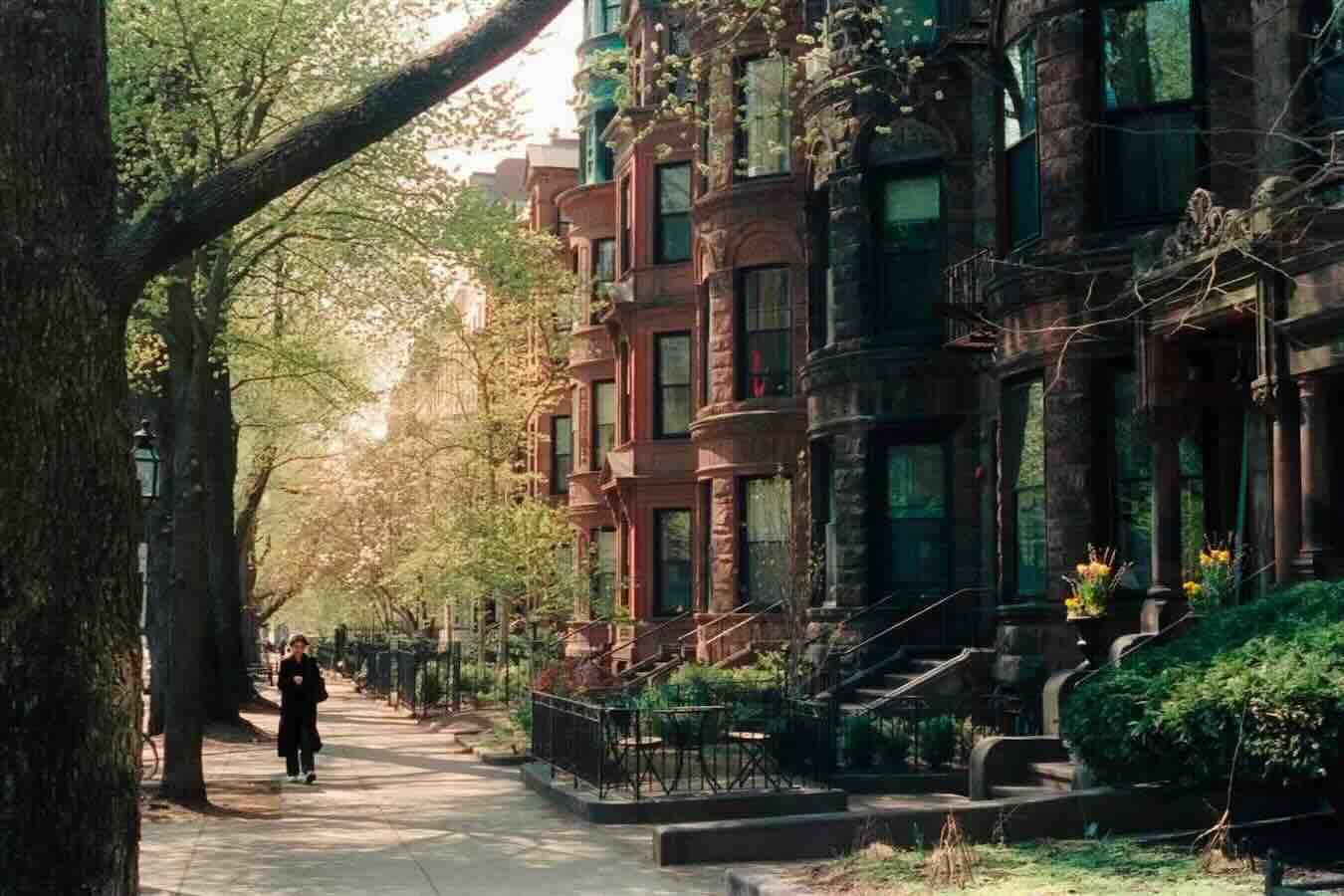 Row of brick townhouses with trees lining a sidewalk; person walks along the path.