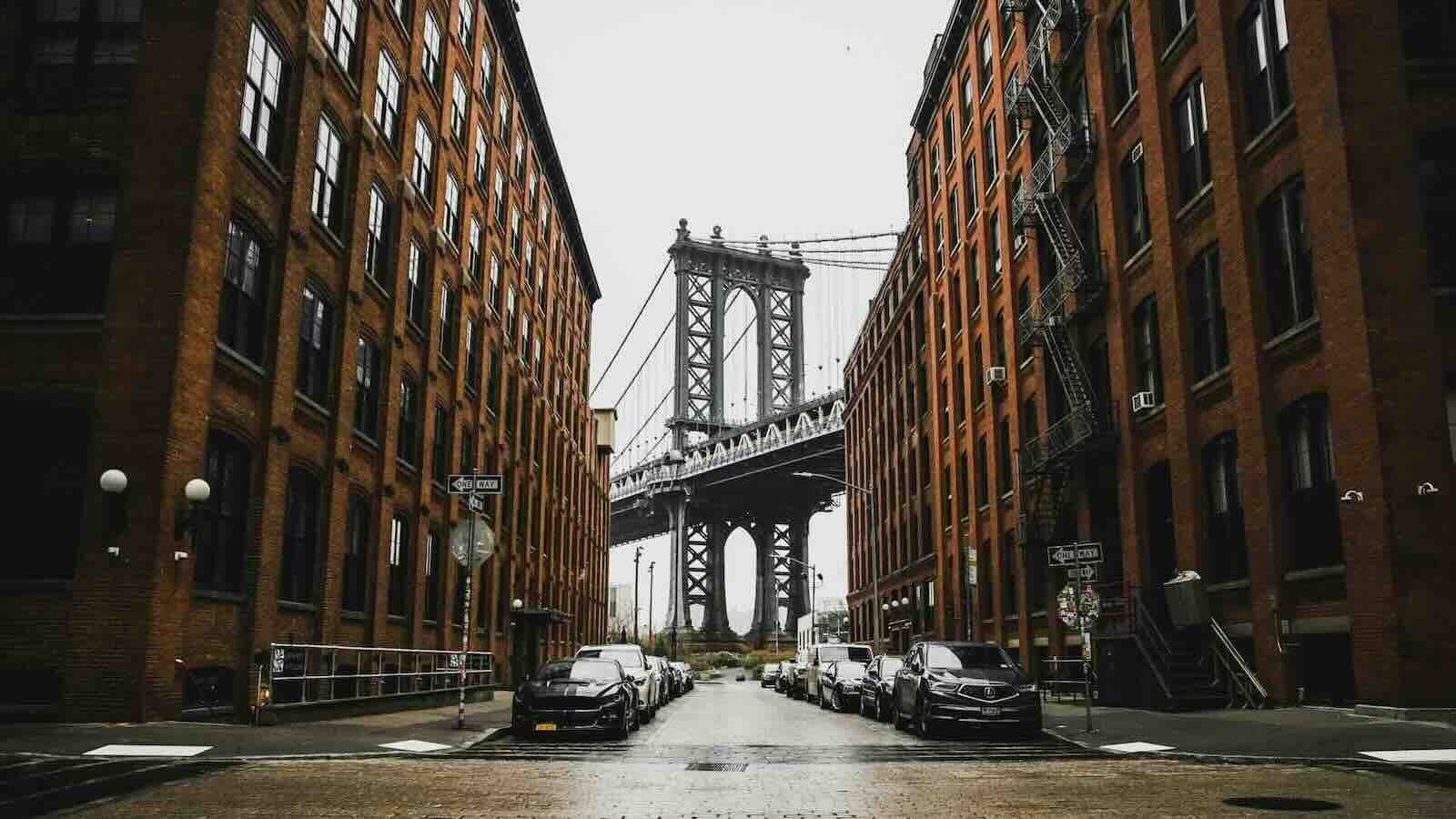 Brick buildings flank a street leading to the Manhattan Bridge. Cars line the street. Overcast sky.