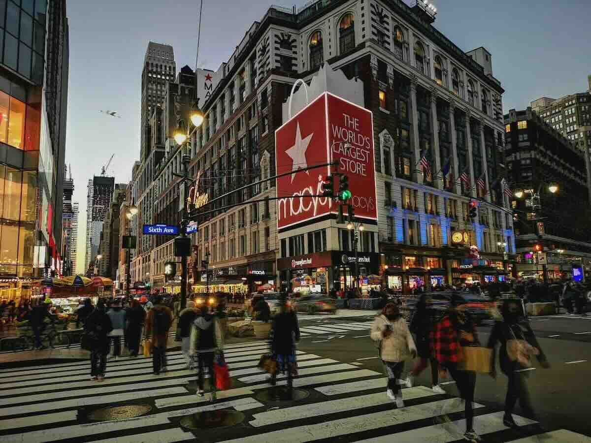People crossing a busy street in front of a tall building with a large red sign.