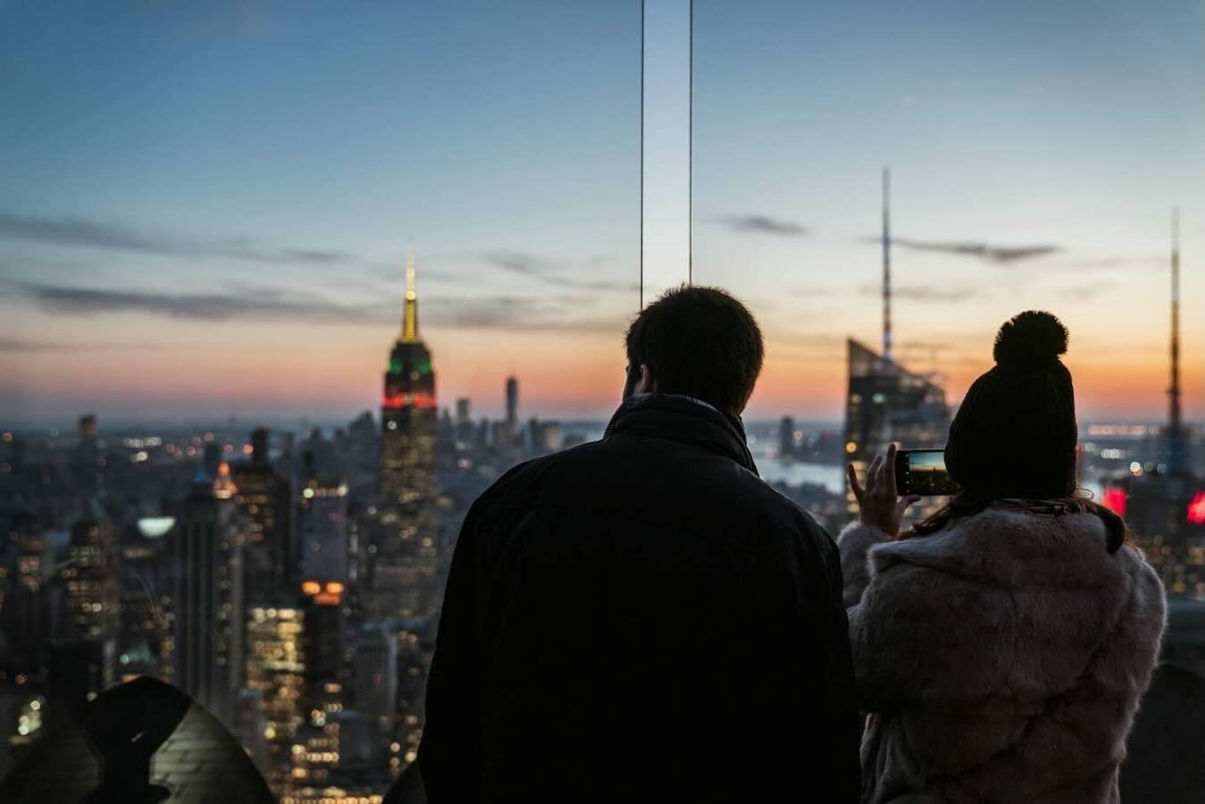 Couple viewing NYC skyline at dusk, woman taking photo, Empire State Building lit up.