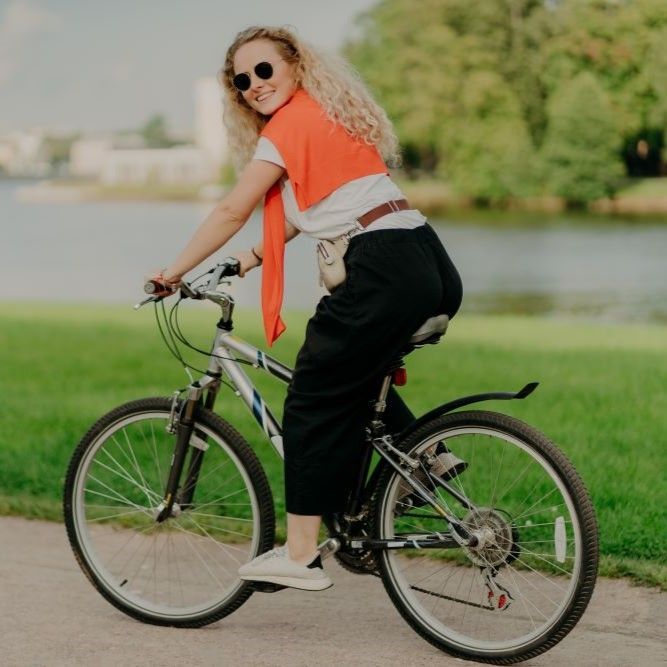 Woman riding a bicycle on a path, smiling. Wearing sunglasses, orange sweater, black pants, and white shoes. Green background.