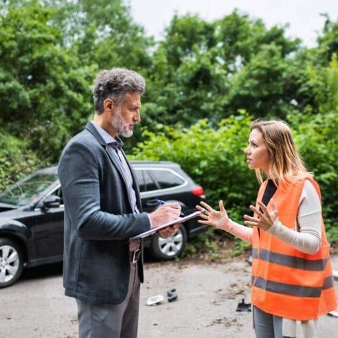 Man in suit taking notes, woman in safety vest gesturing; car accident scene.