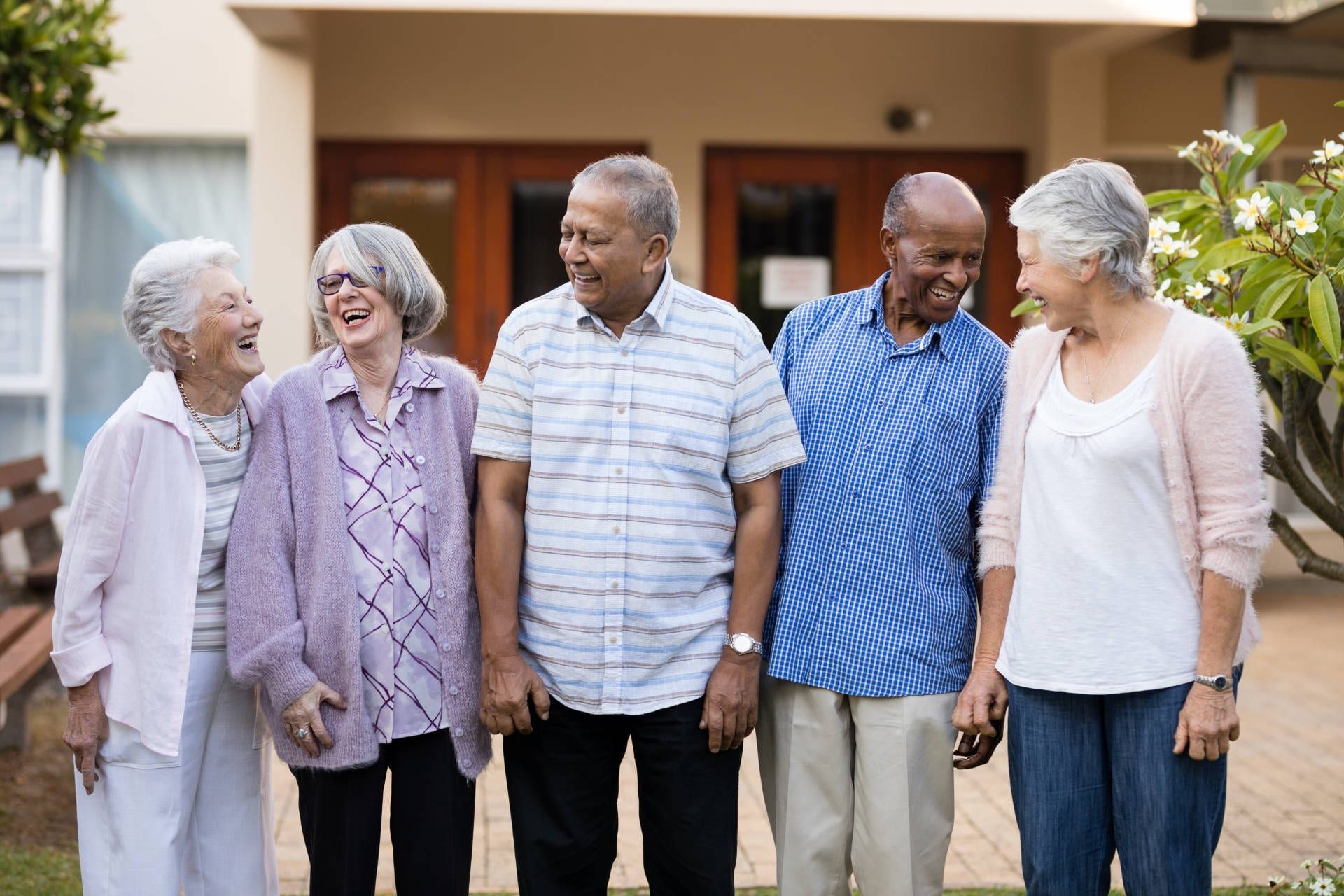 Five older adults smiling and laughing outdoors; in front of a building, with greenery in background.