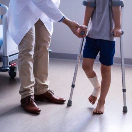 Doctor helping a child walk with crutches; child's leg wrapped in bandage. Indoors.
