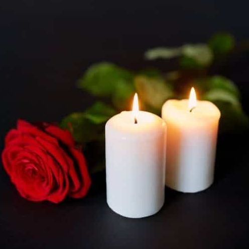 Two lit white candles and a red rose on a dark background.