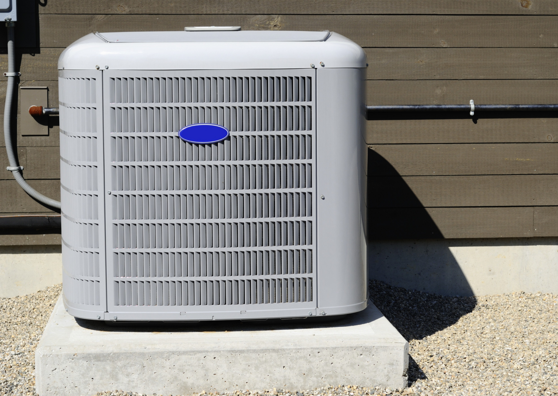Gray air conditioner on a concrete pad against a wooden wall with pebbles.