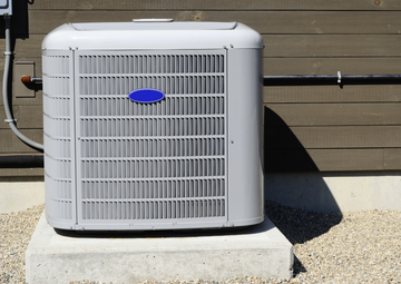 Gray air conditioner on a concrete pad against a wooden wall with pebbles.