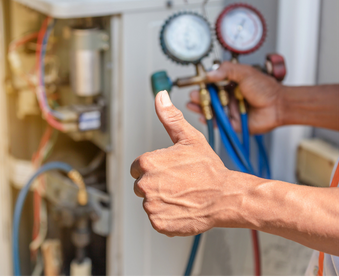 A repair technician with a thumbs up, holding gauges, working on an air conditioning unit.