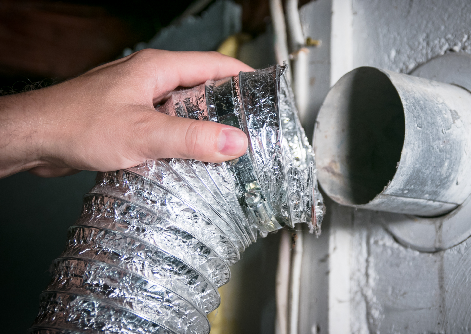A person's hand disconnecting a silver dryer vent hose from a wall vent.