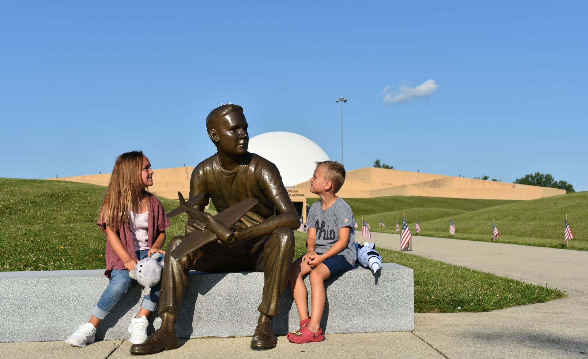 Two children are sitting next to a statue of Neil Armstrong outside of the Armstrong Air & Space Museum.