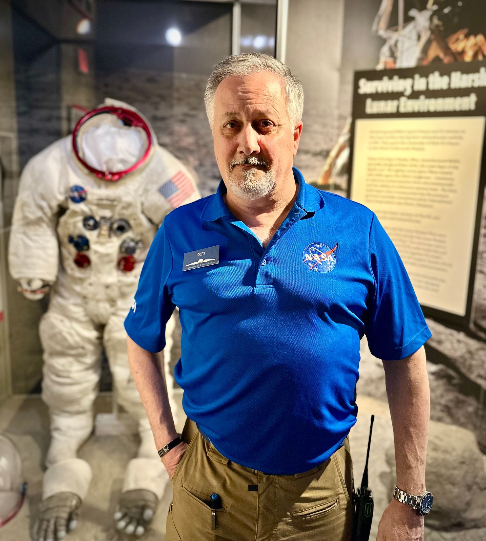 Greg Brown standing in front of the Apollo spacesuit wearing a blue NASA polo and cargo pants.