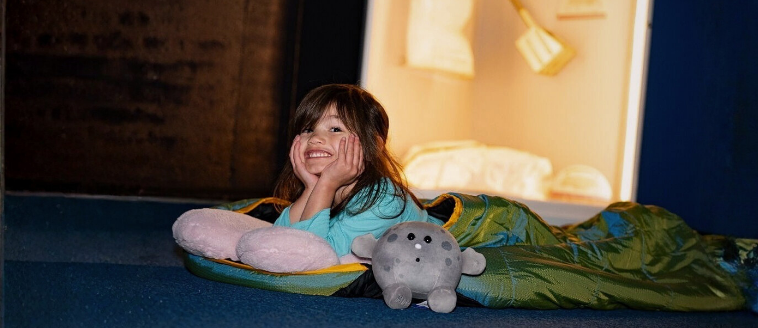 A child in a sleeping bag smiling as she sleeps underneath a Moon rock in one of the museum's galleries.