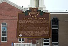 A historical marker for Dudley Nichols is in front of a brick building.