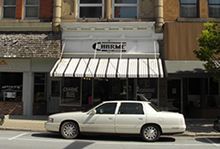 A white car is parked in front of the Rhine and Brading Drug Store with an awning.