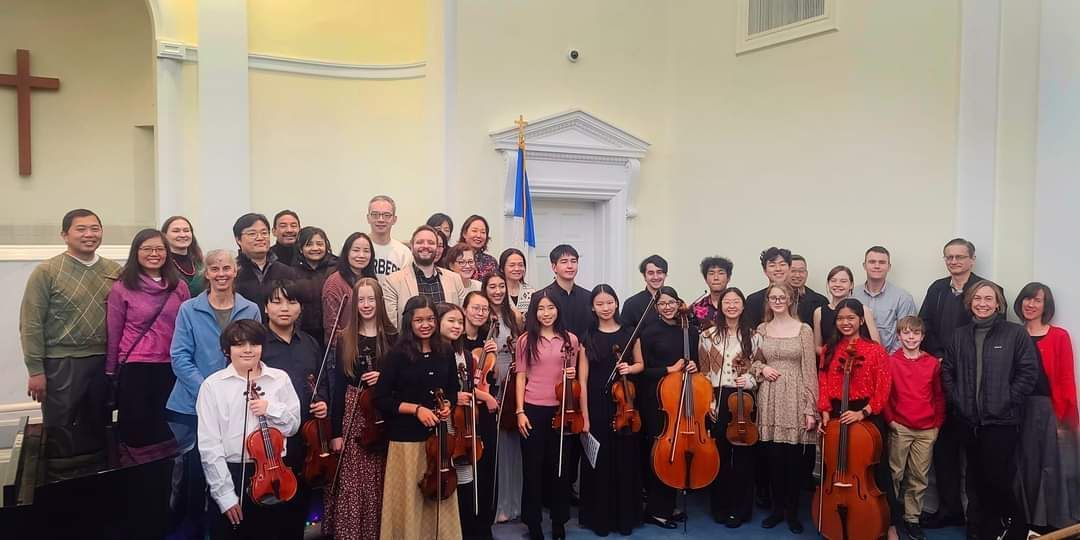 A large group of Koart Academy students standing next to each other holding violins and cello on a stage in First Baptist Church.