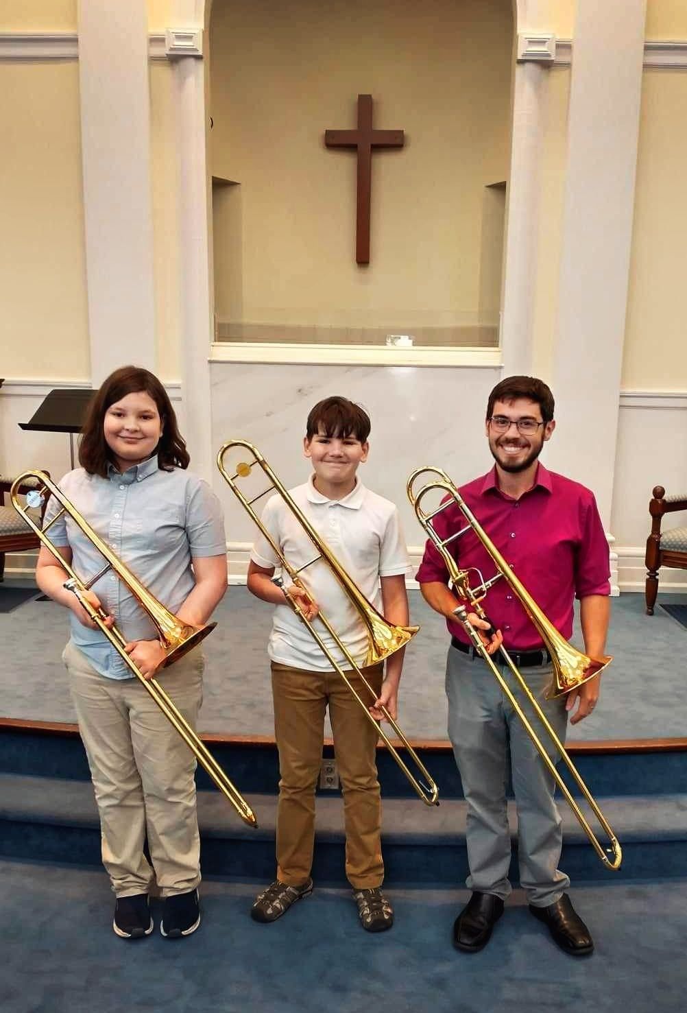 Three children are holding trombones in front of a cross in First Baptist Church.