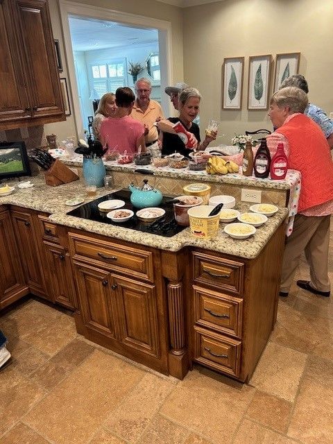 Older adults gather in a kitchen, serving ice cream from a countertop with bowls, sauces, and toppings.