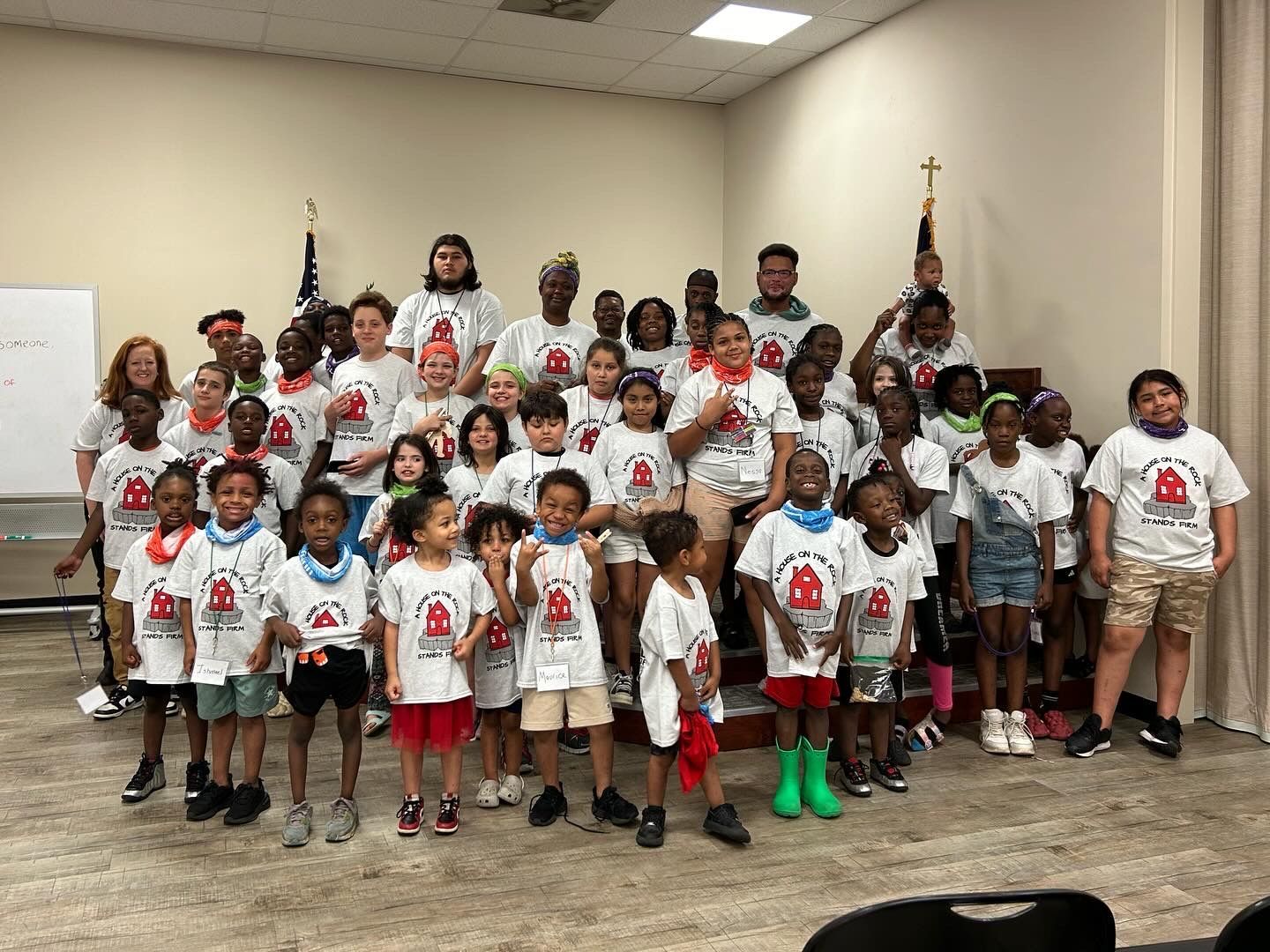 A large group of children in matching summer camp shirts are posing for a picture in a room.