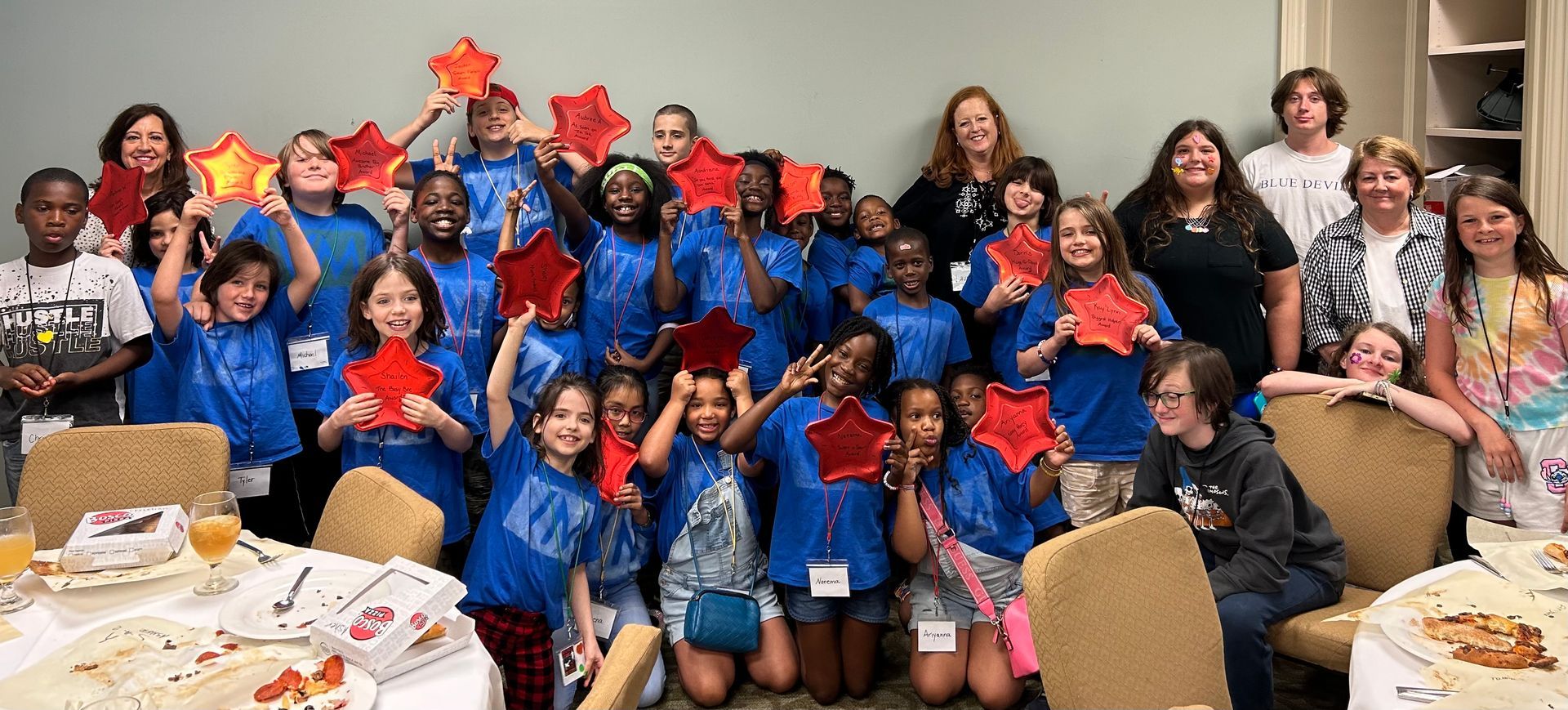 A group of children in matching summer camp shirts are posing for a picture, holding red stars.