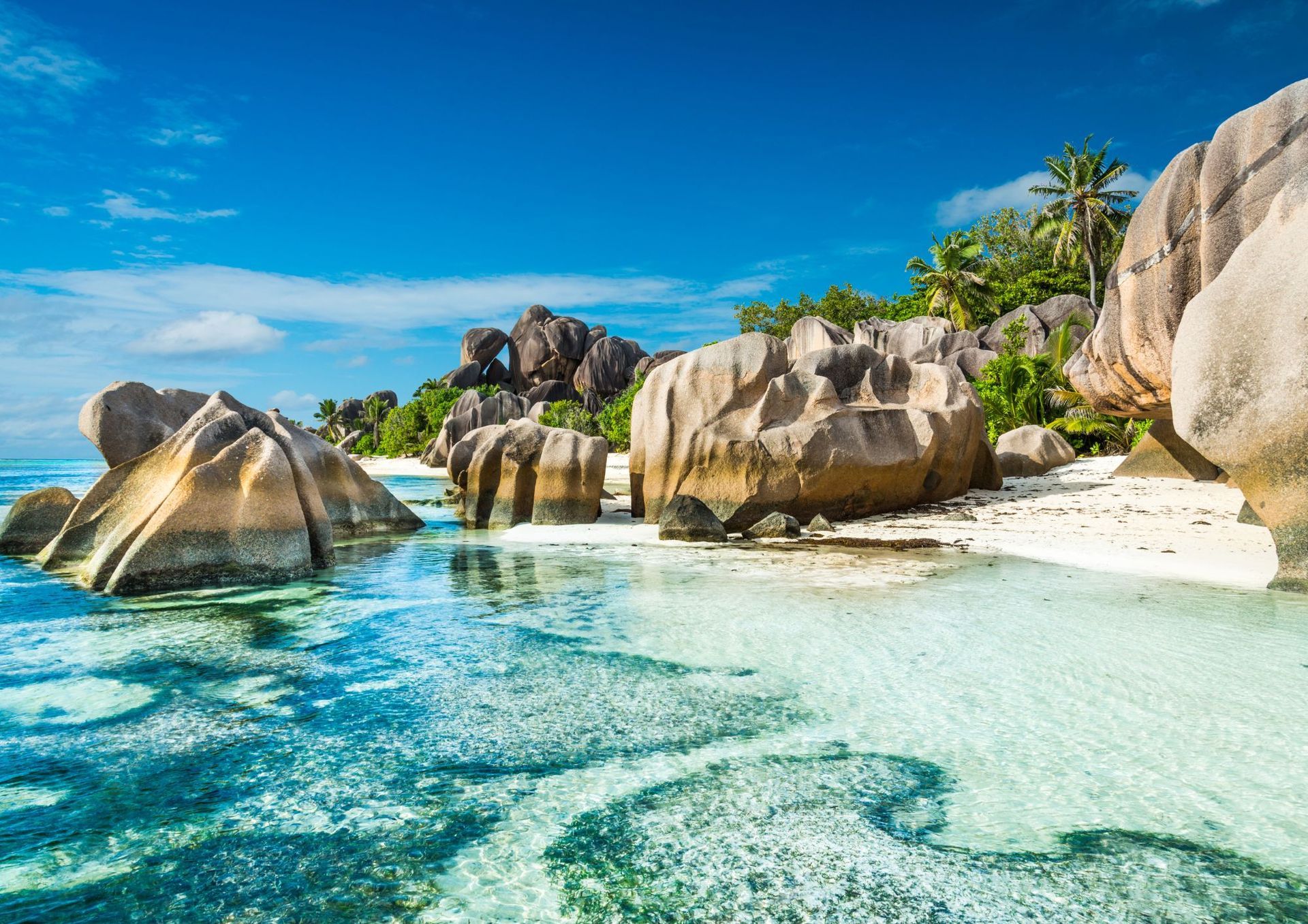 Beach with large granite boulders, clear turquoise water, white sand, and blue sky.