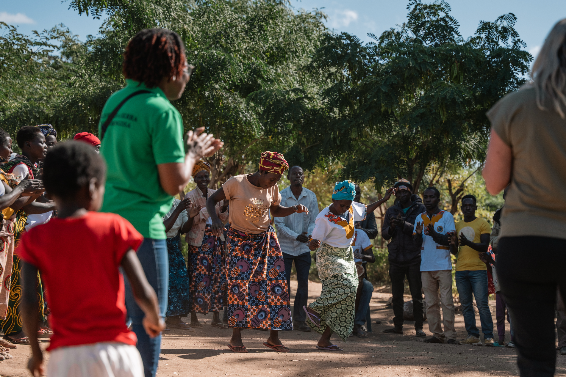 People dancing outdoors, surrounded by onlookers.  A woman in green watches.  Bright, sunny day.