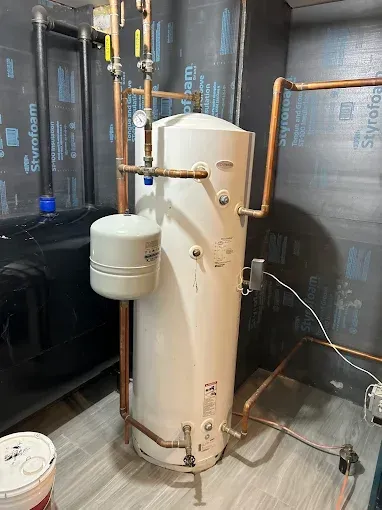 A tall, white water heater with copper pipes in a room with black insulation and gray flooring.