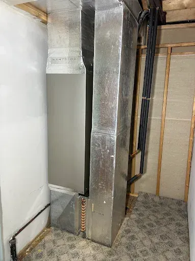 HVAC system in a small room with silver ductwork, pipes, and electrical conduit against white and wood walls.