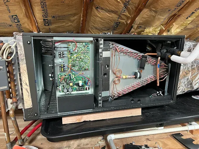 Open air conditioning unit in an attic, revealing electrical components and copper coils.