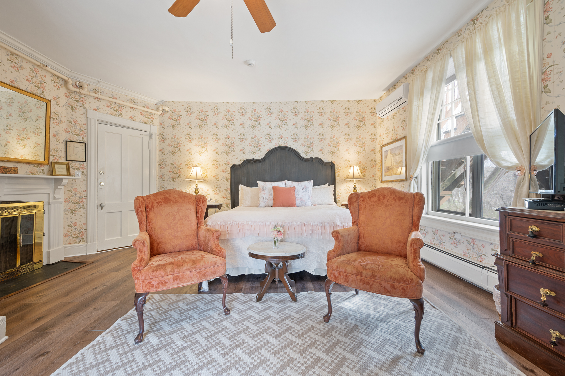 A bedroom with a large bed , two chairs , a fireplace and a ceiling fan.