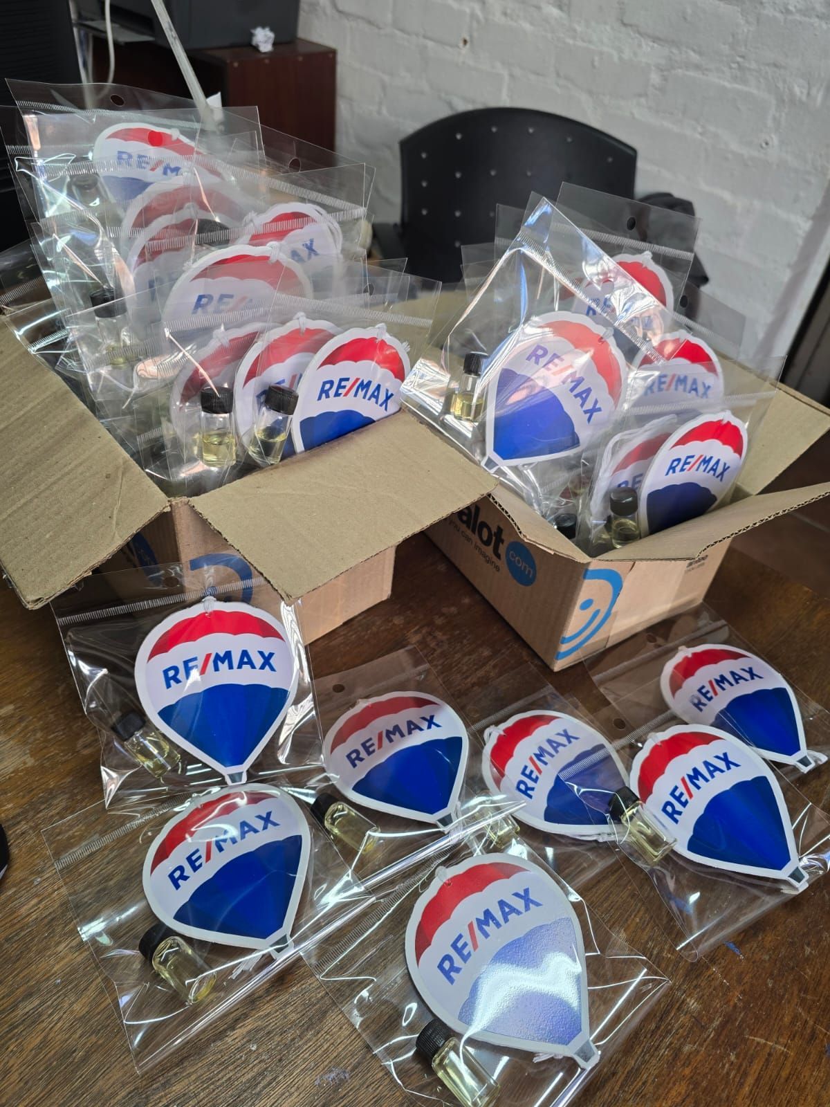 RE/MAX logo keychains, packaged in clear bags, spilling out of a cardboard box on a wooden table.