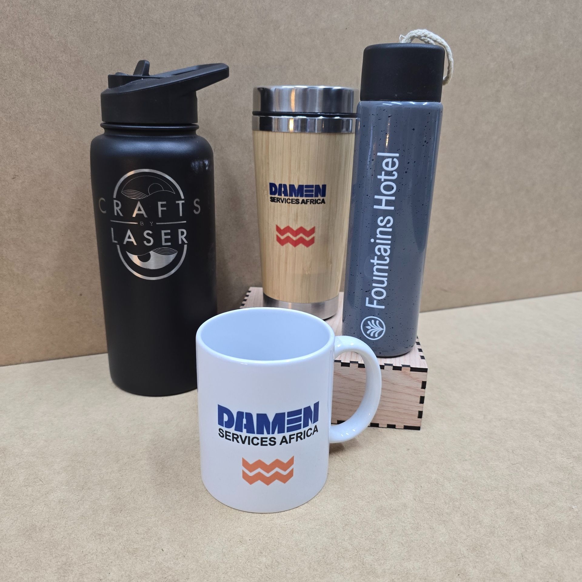 Promotional drinkware: black water bottle, bamboo travel mug, tall gray thermos, and a white mug. All branded with logos.