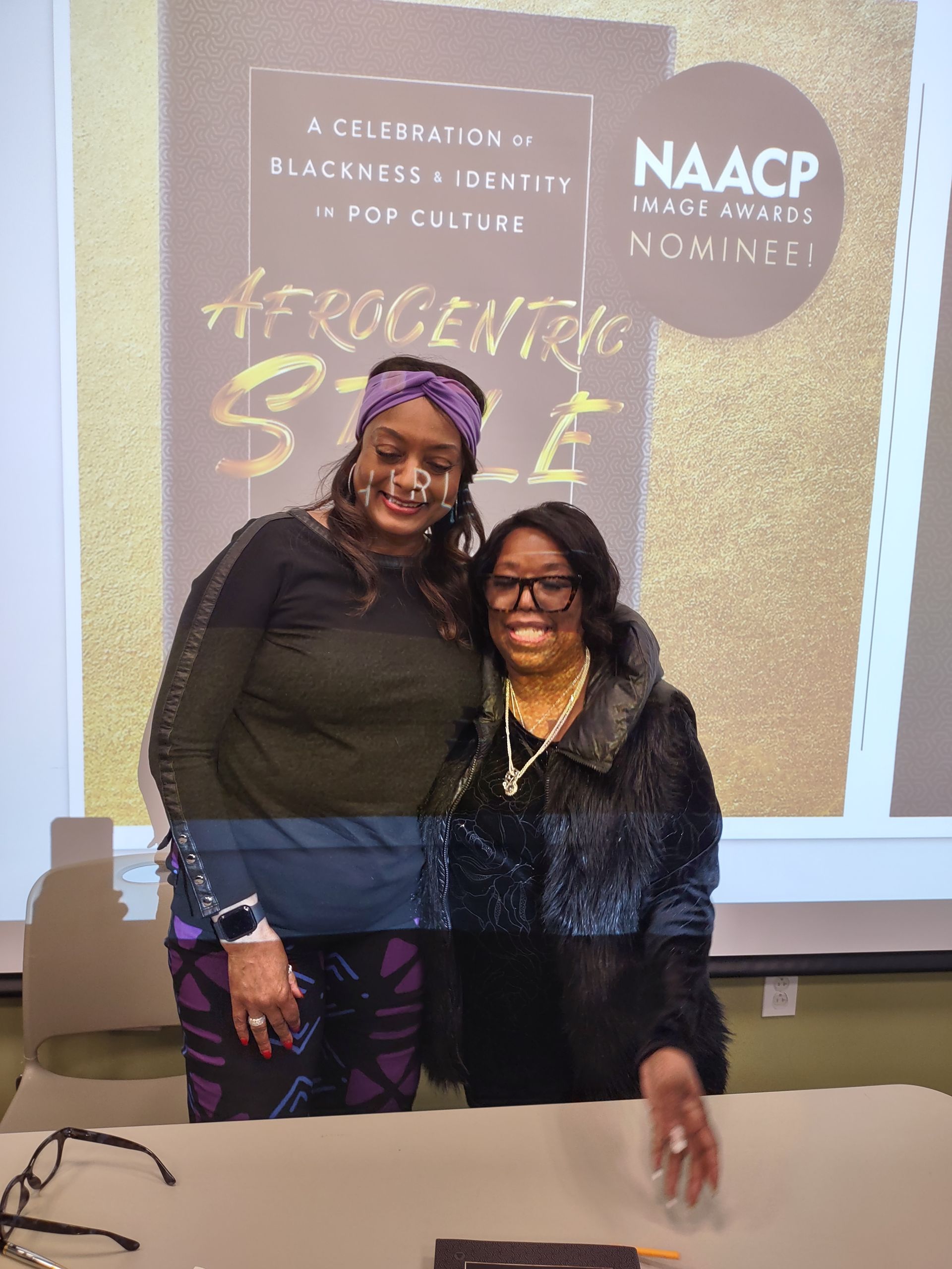 Two women are posing for a picture in front of a projector screen.