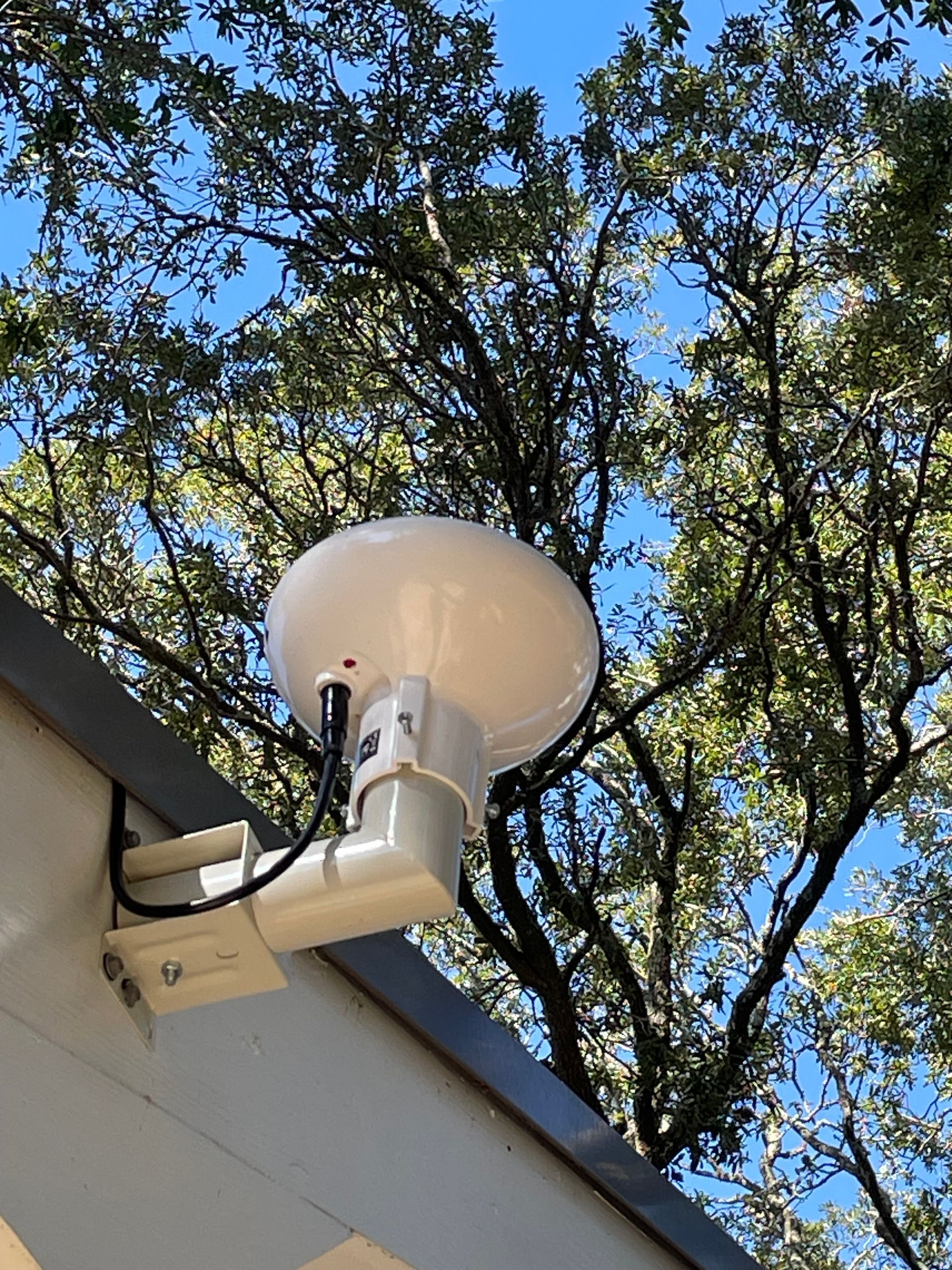 White satellite dish mounted on a building corner, trees in the background, blue sky.