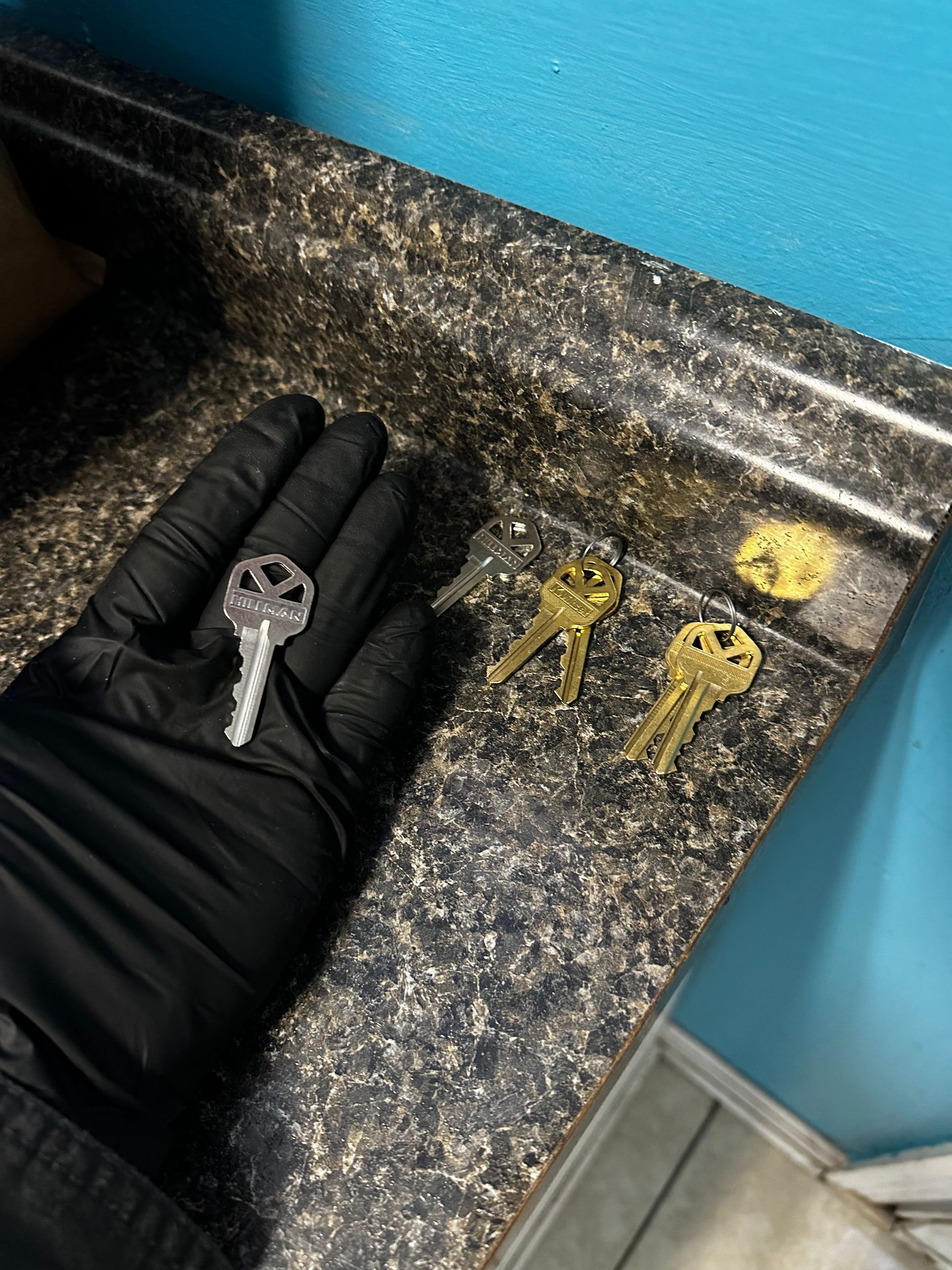 A gloved hand holds a silver key; three gold keys sit on a speckled countertop near a blue wall.