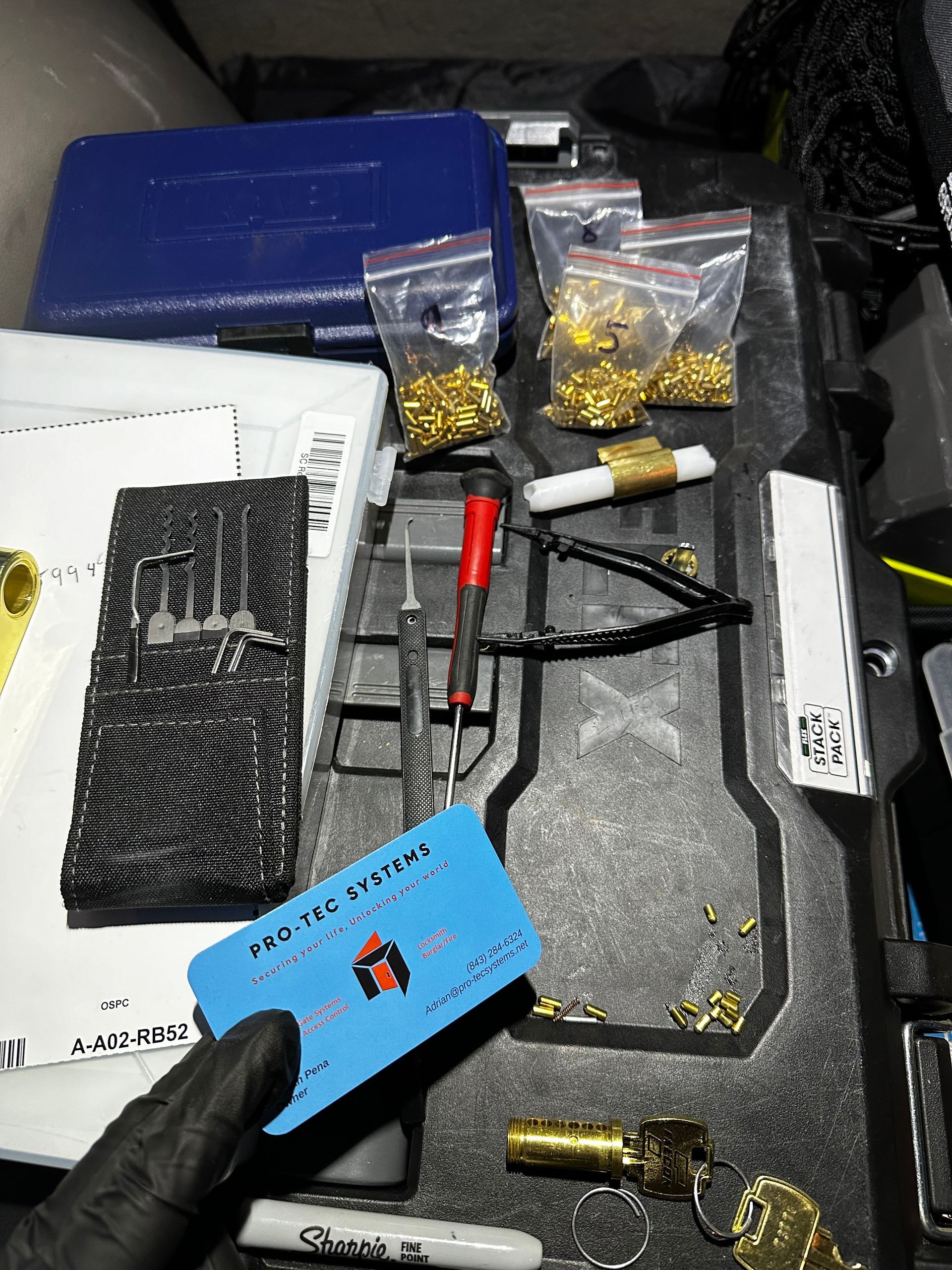 Close-up of locksmith tools, including pins, picks, and a key. A hand in a black glove holds a blue card.