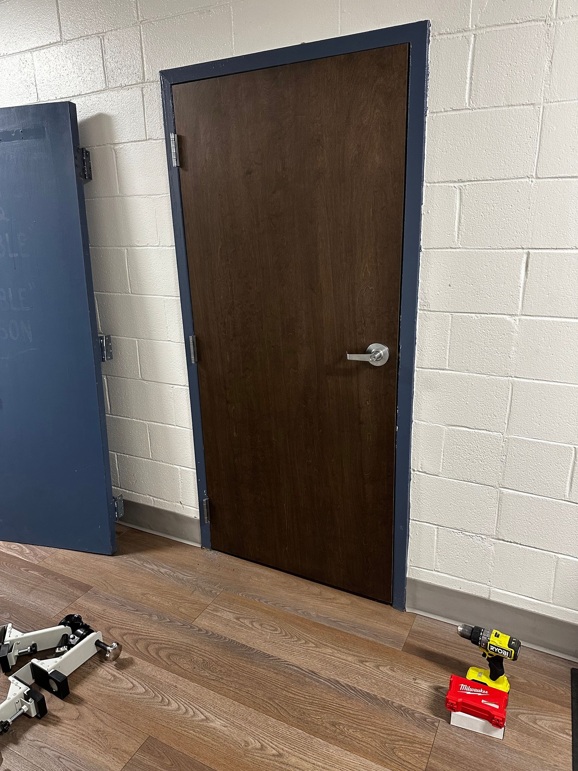 Brown door with blue trim in a white brick room. A blue door is propped on the wall, and a drill is on the floor.