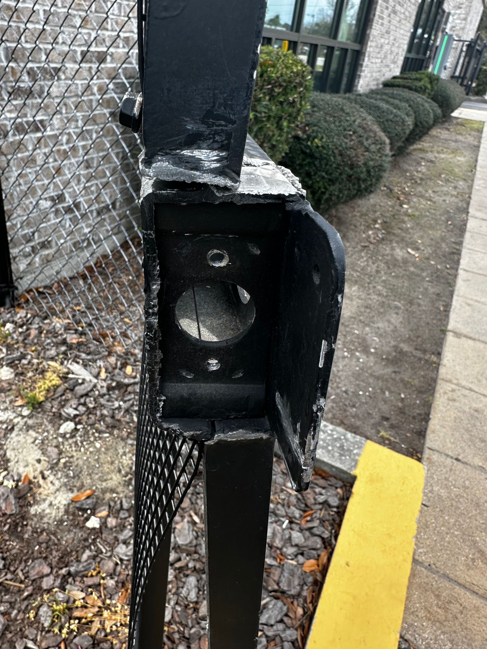 Black metal fence post with a round hole, likely for a light or speaker, next to a sidewalk and bushes.