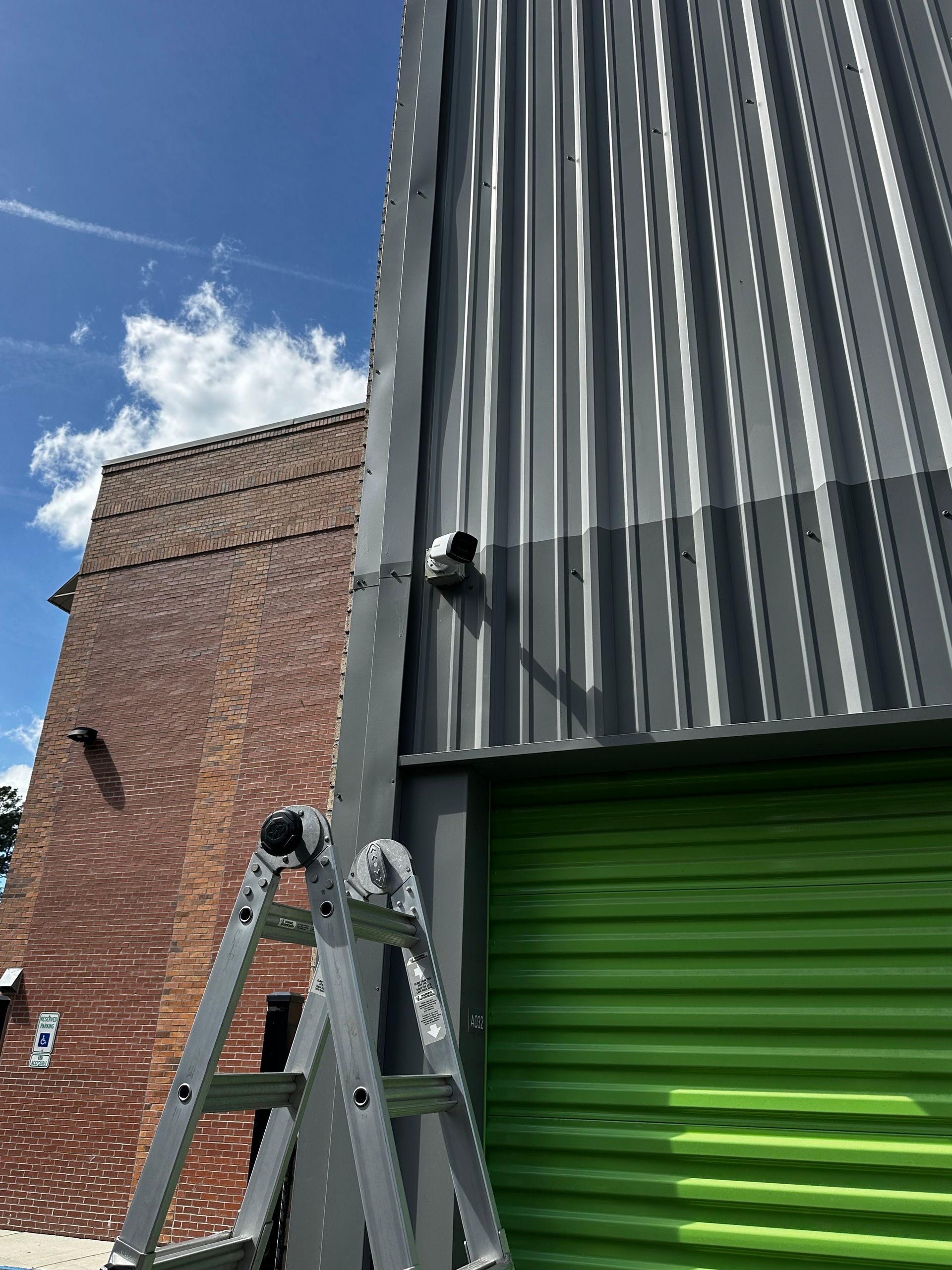 Ladder next to gray corrugated building; green door below, brick building to the left, blue sky.