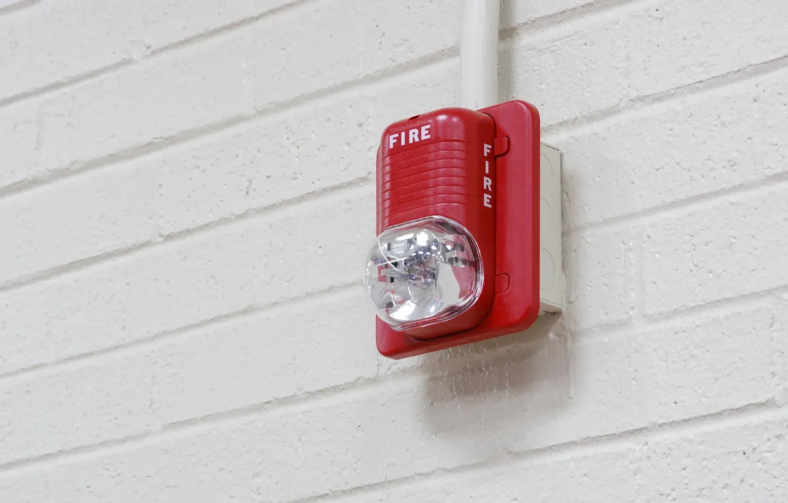 Red fire alarm with strobe light mounted on a white brick wall.