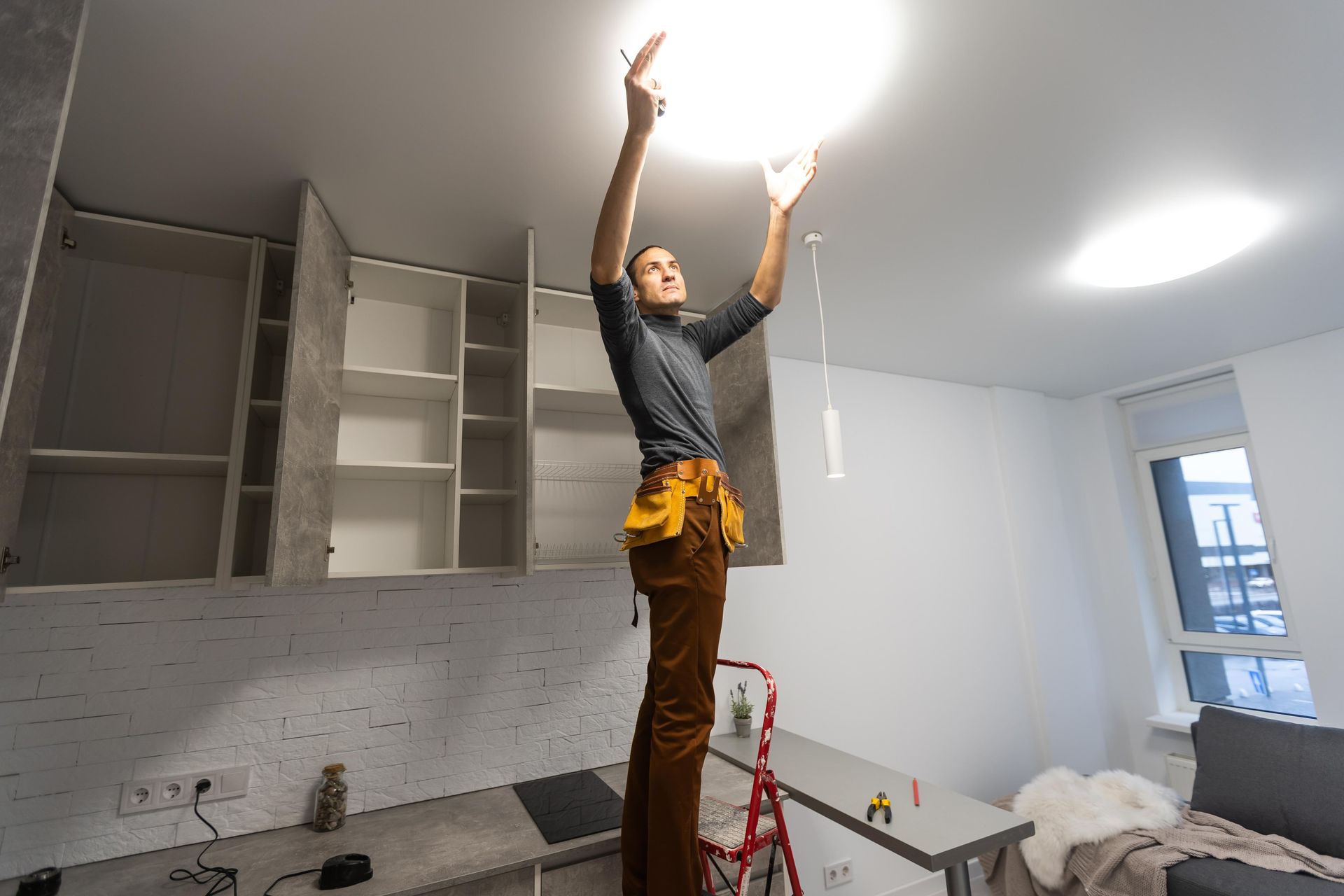 Person on a stepladder installing a ceiling light in a room with cabinets and a table.