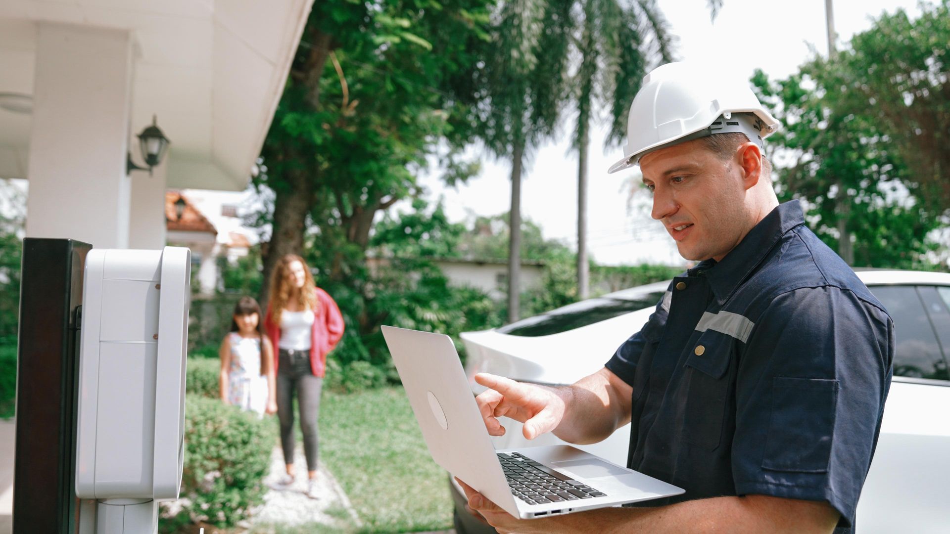 Man in hard hat using laptop near charging station, two people behind him.